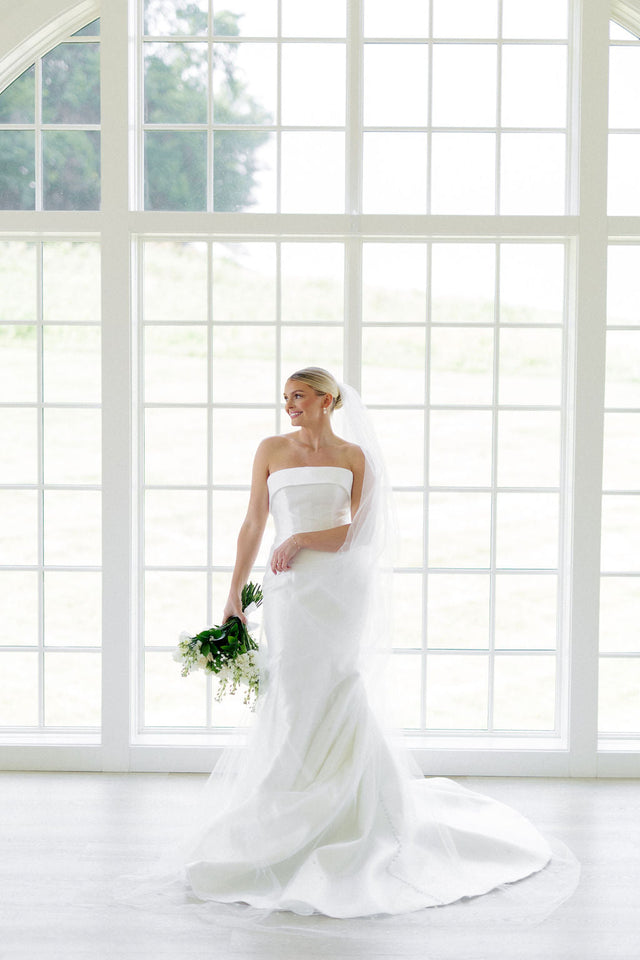 Bride in a white gown holding a bouquet by tall windows