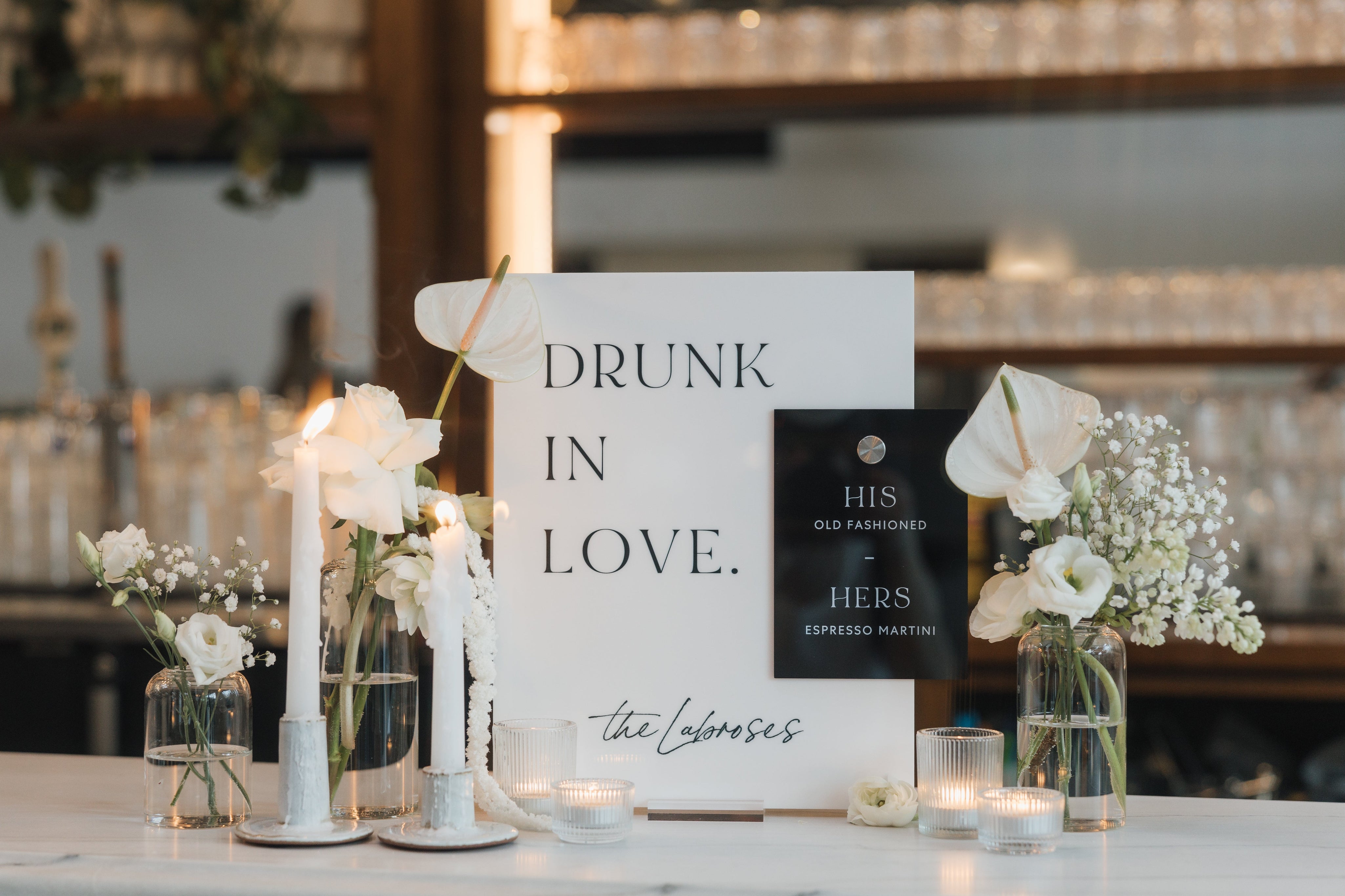 Elegant wedding bar setup with 'Drunk in Love' acrylic sign and signature drink menu cards surrounded by white floral arrangements and candles