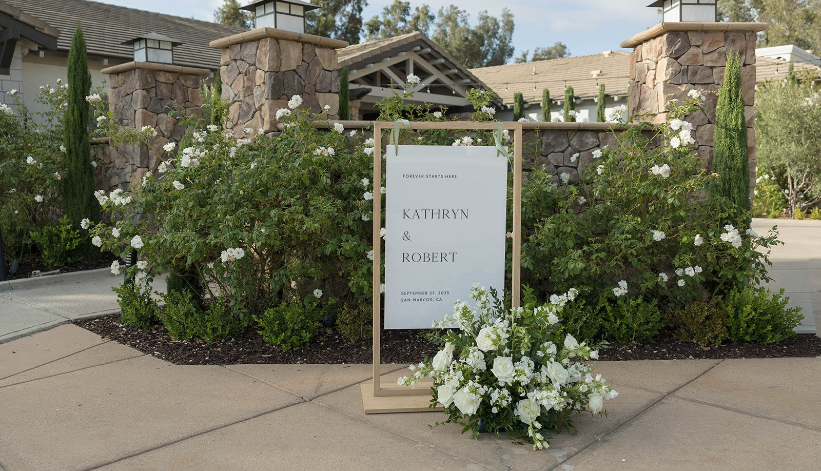 Custom wedding welcome sign for Kathryn and Robert on September 27, 2025 in San Marcos, CA displayed on a wooden easel with white floral arrangements at the entrance of a stone venue