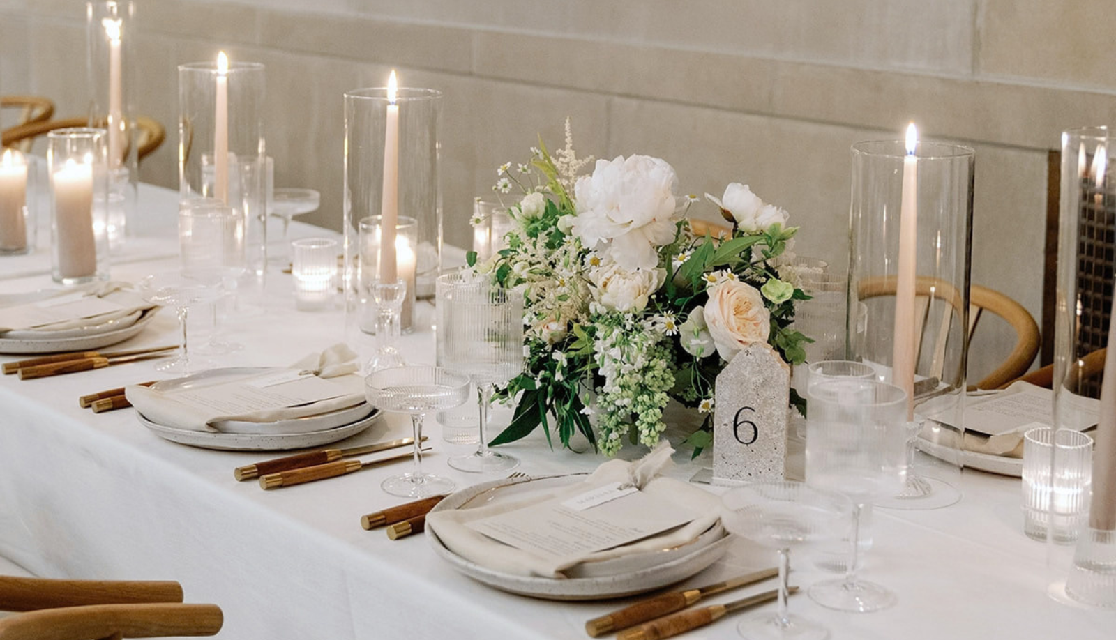 Elegant wedding reception table setting with white linens, stone table number 6, tall lit candles in glass holders, and a lush white and green floral centerpiece
