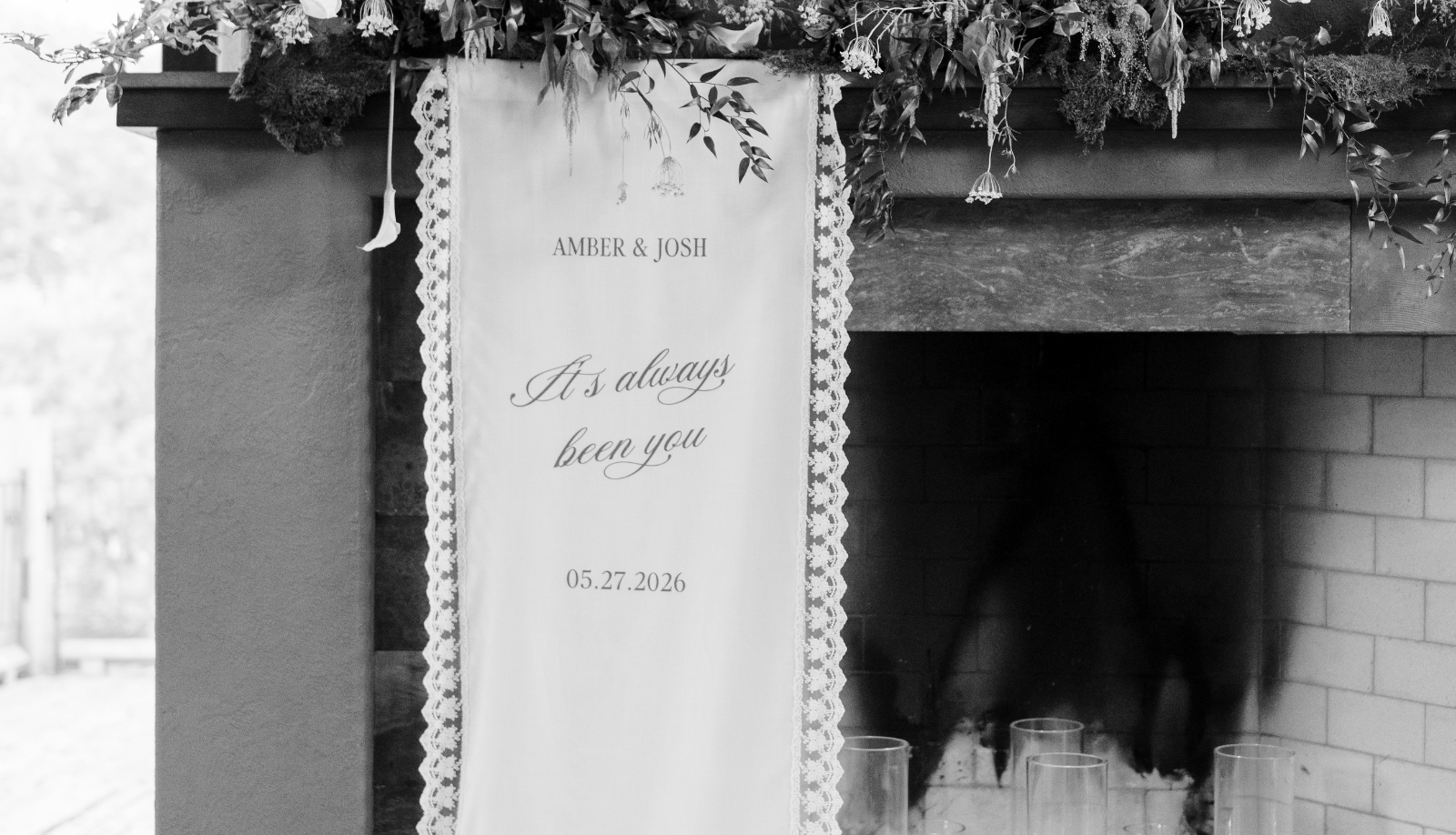 Custom wedding welcome sign for Amber and Josh reading 'It's always been you' with the date 05.27.2026, displayed with lace trim and floral greenery above a fireplace