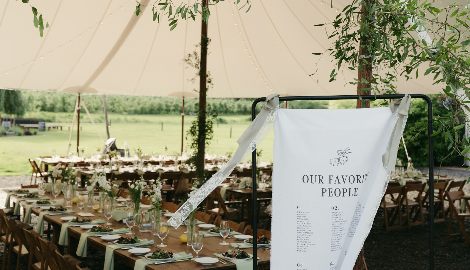 Custom linen wedding seating chart titled 'Our Favorite People' displayed on a black stand at an outdoor tented wedding reception with long wooden tables, floral centerpieces, and string lights