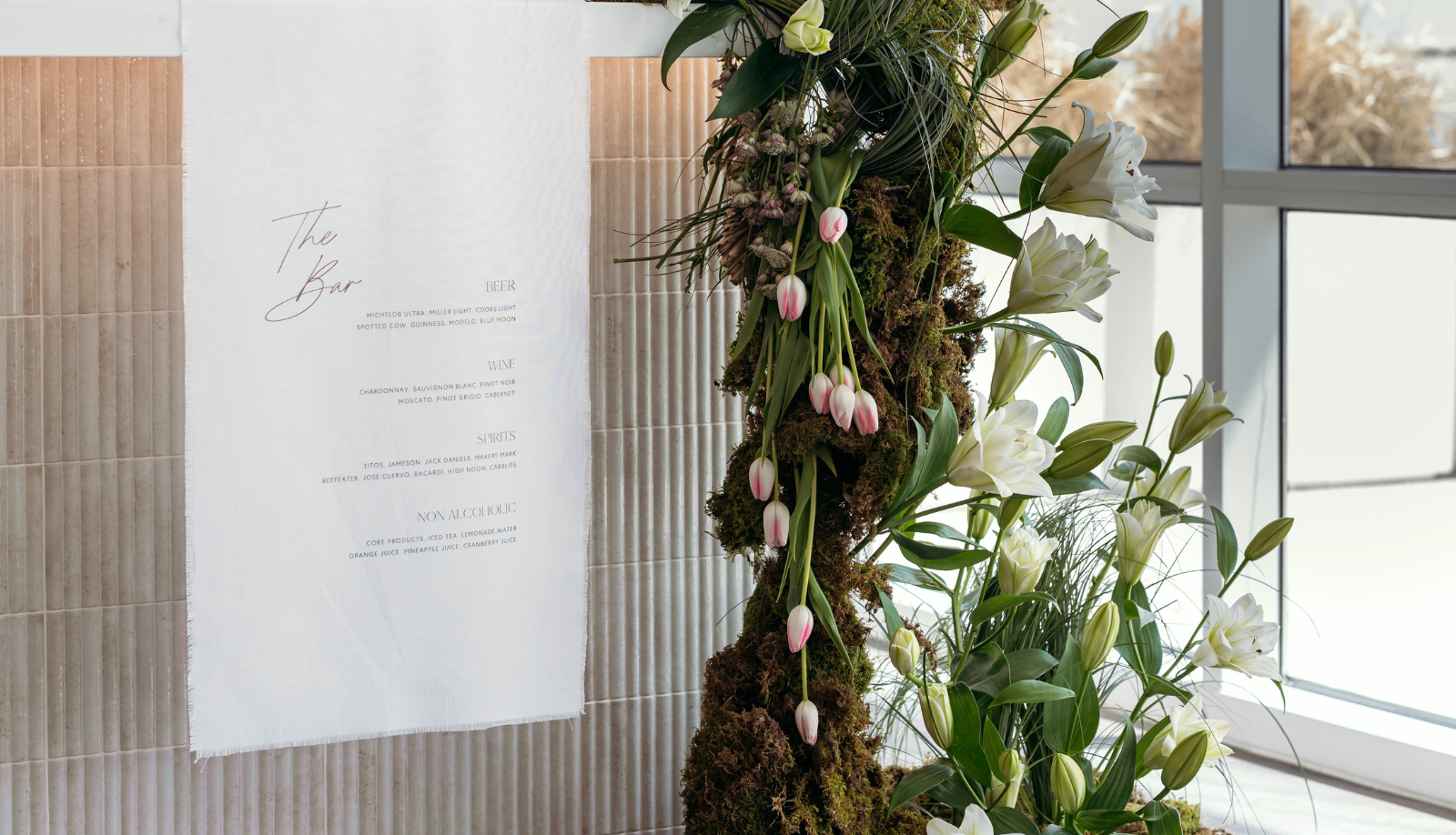 Custom linen wedding bar menu sign titled 'The Bar' displayed with a lush floral and moss arrangement featuring white tulips and lilies at an elegant reception venue