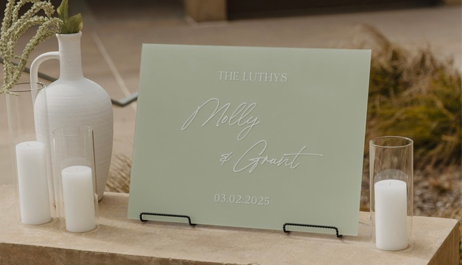 Custom sage green acrylic wedding welcome sign for Molly and Grant dated March 2, 2025, displayed on a black stand with white candles and a white vase on a stone surface